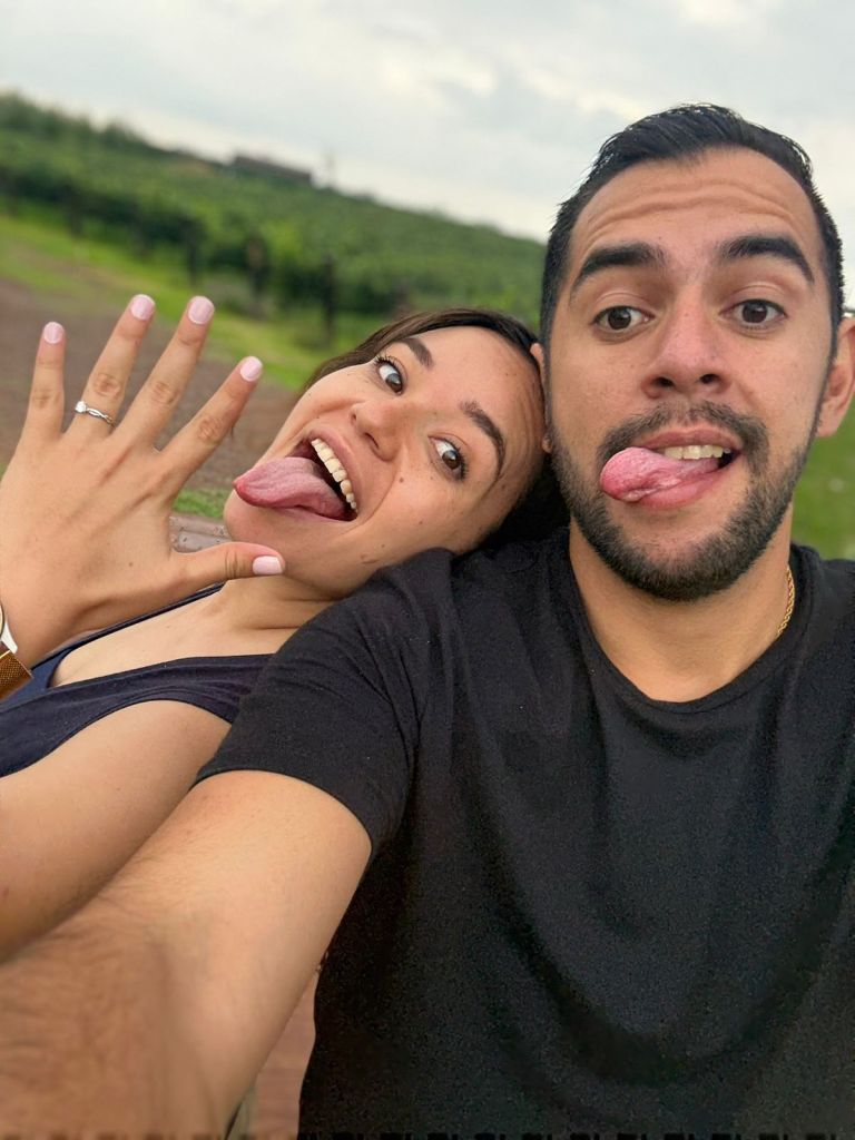 A man and a woman tounge out, facing the camera and posing in a selfie-like setting, possibly outdoors. Being unable to co...
