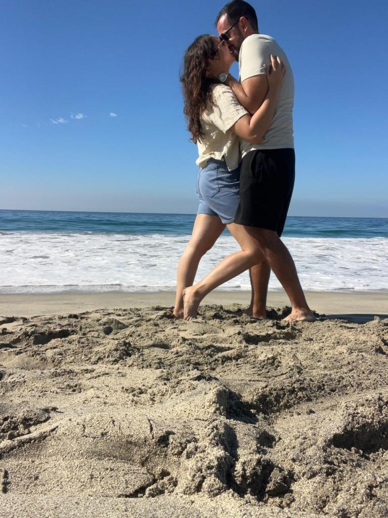 The image shows a romantic scene of a couple embracing on a sandy beach, with the ocean waves and clear blue sky in the ba...