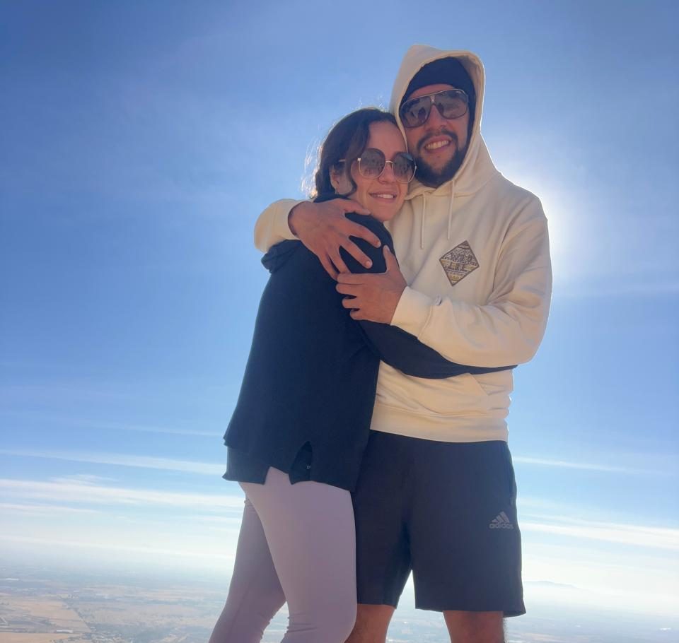 The image depicts a man and a woman standing on a rocky outcrop, overlooking a vast landscape below them. The woman is wea...